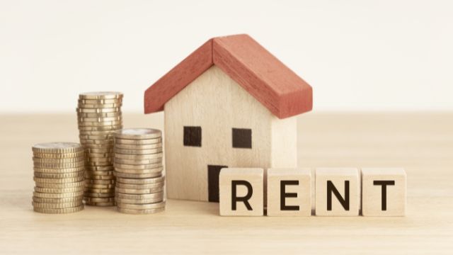 Stacks of coins next to a small wooden house model spelling out RENT